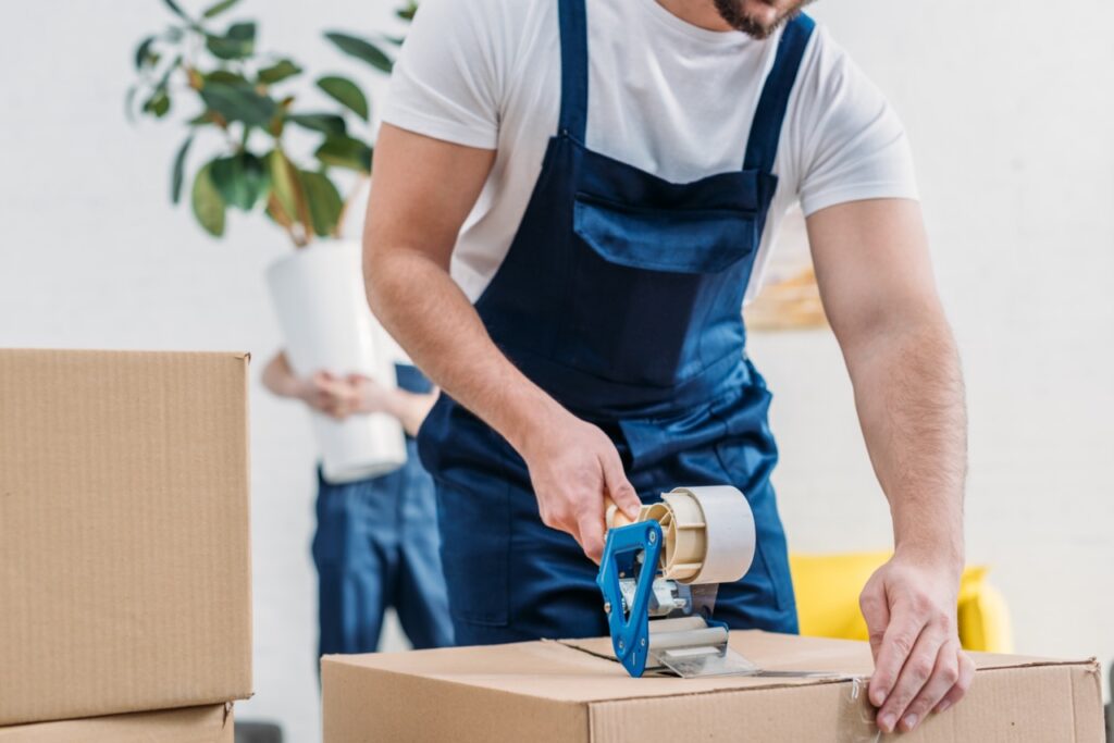 Mover sealing a box with tape while another carries a potted plant